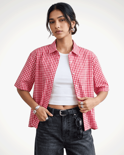 A model wearing Red Madras Check Shirt boxy fit shirt by Covert over a white crop top, paired with dark gray jeans and a black belt, against a plain white studio background.